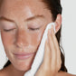 Woman cleaning her face with a white towel against a neutral background