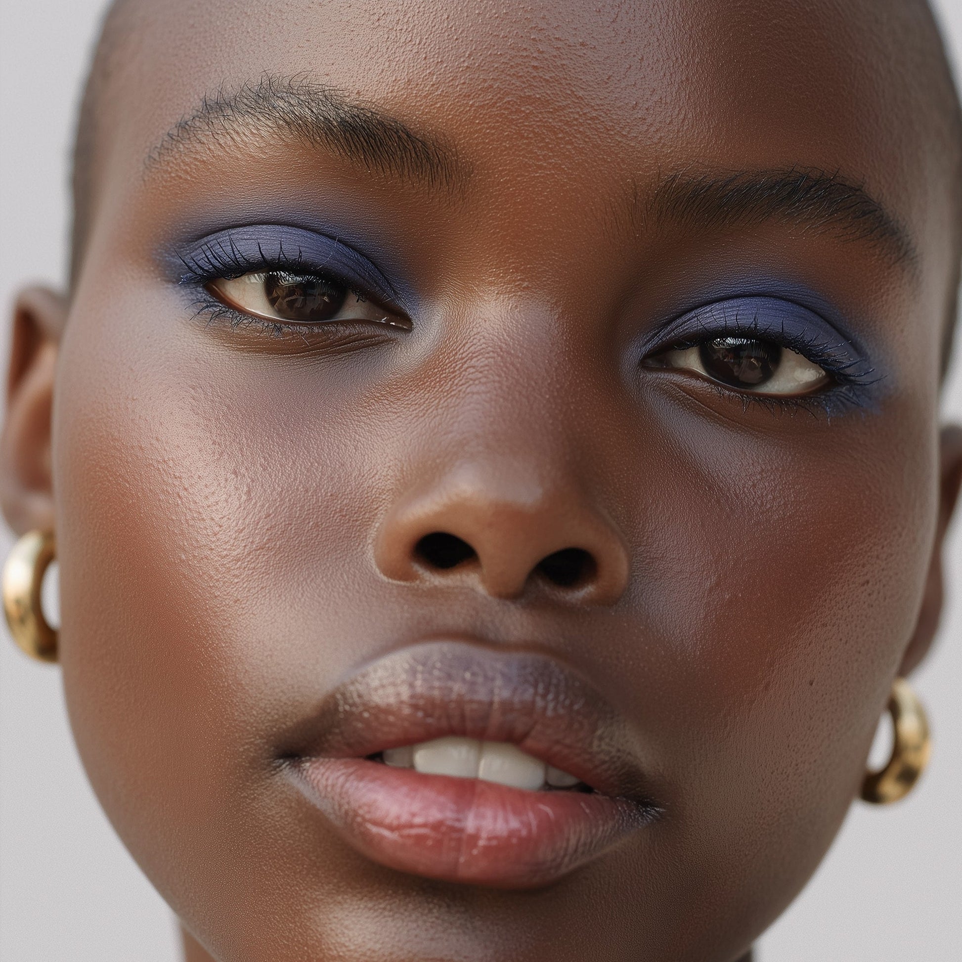 Close-up of a woman's face with blue eyeshadow and gold earrings on a neutral background (Blue)