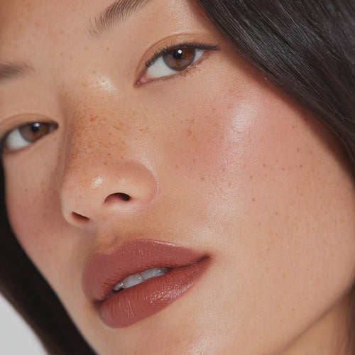Close-up of a person’s face featuring soft natural makeup, glossy nude lips, and visible freckles.