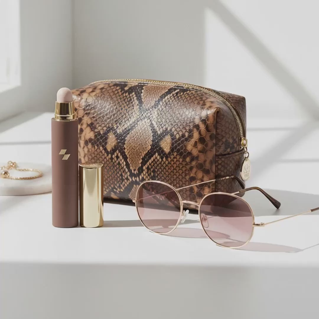 Tinted lip balm stick standing beside a snakeskin-patterned cosmetic bag and round sunglasses on a white surface.