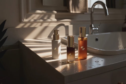 Three skincare products in minimalist bottles displayed on a bathroom counter next to a sink, lit by soft natural sunlight.